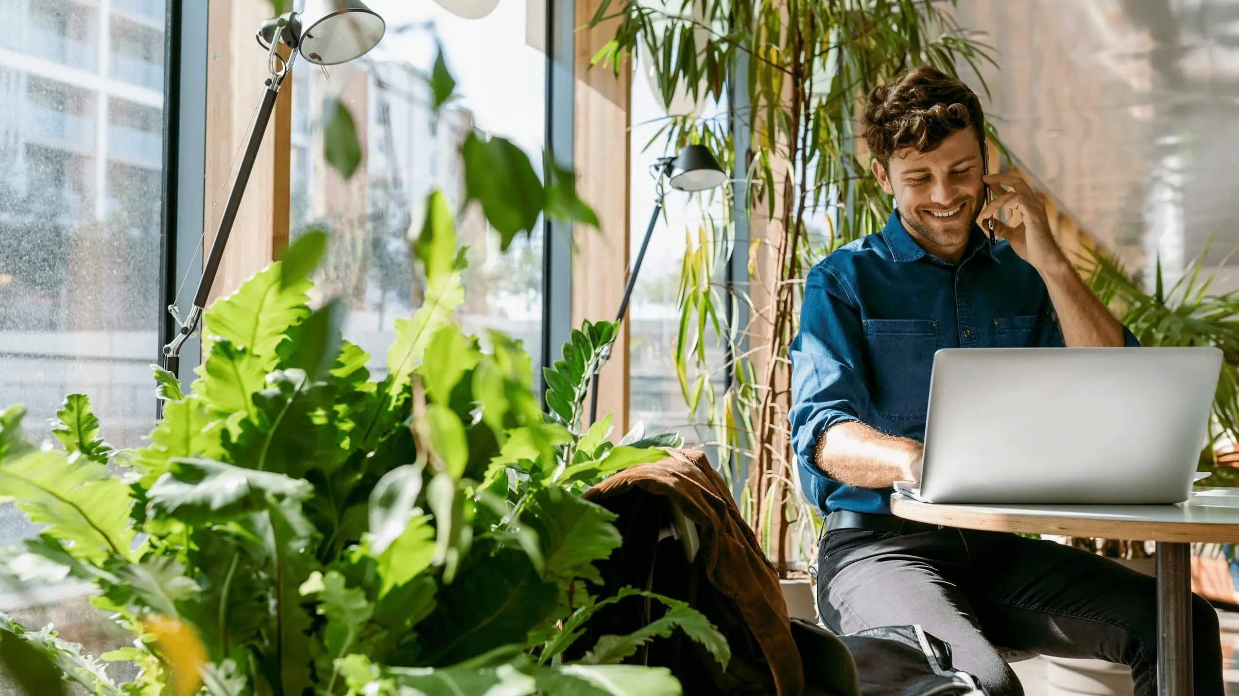 Mann sitzt am Telefon vor einem Laptop, es sind viele Pflanzen um ihn herum, als Symbol f&uuml;r Klima
