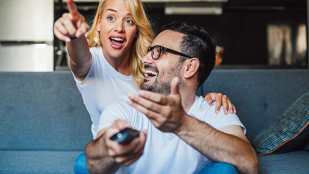Ein Paar sitzt gemeinsam auf der Couch, der Mann hat eine Fernbedienung in den H&auml;nden