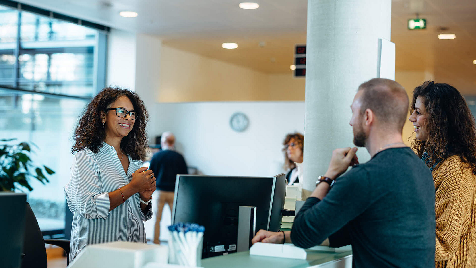 Empfangsmitarbeiterin begr&uuml;&szlig;t zwei Personen am Tresen
