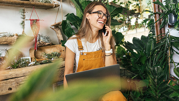 Eine Frau sitzt von Pflanzen umgeben vor einem Laptop und telefoniert