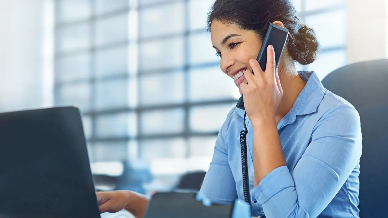 Zu sehen ist eine l&auml;chelnde Frau mit einem Telefonh&ouml;rer am Ohr. Sie sitzt an Ihrem Arbeitsplatz vor einem Laptop und scheint zu telefonieren.