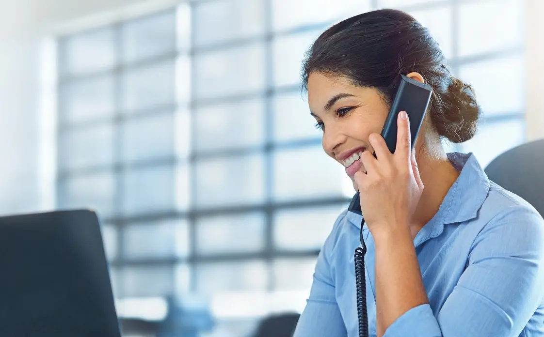 Zu sehen ist eine lächelnde Frau mit einem Telefonhörer am Ohr. Sie sitzt an Ihrem Arbeitsplatz vor einem Laptop und scheint zu telefonieren.