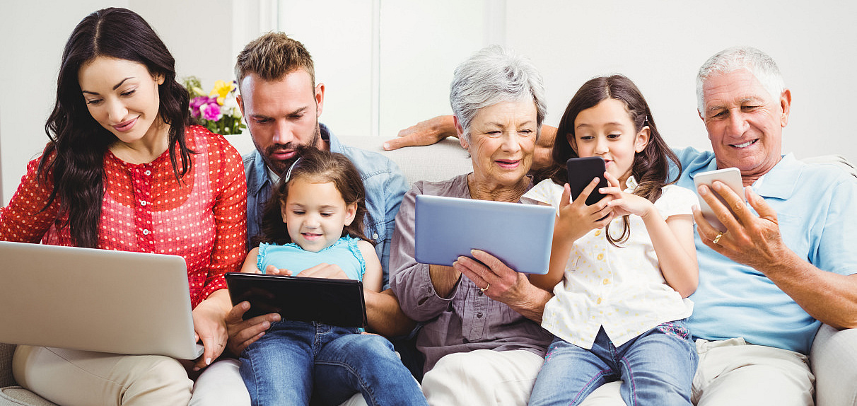 Eine Mehr-Generationen-Familie sitzt gemeinsam auf der Couch vor verschiedenen diditalen Endgeräten