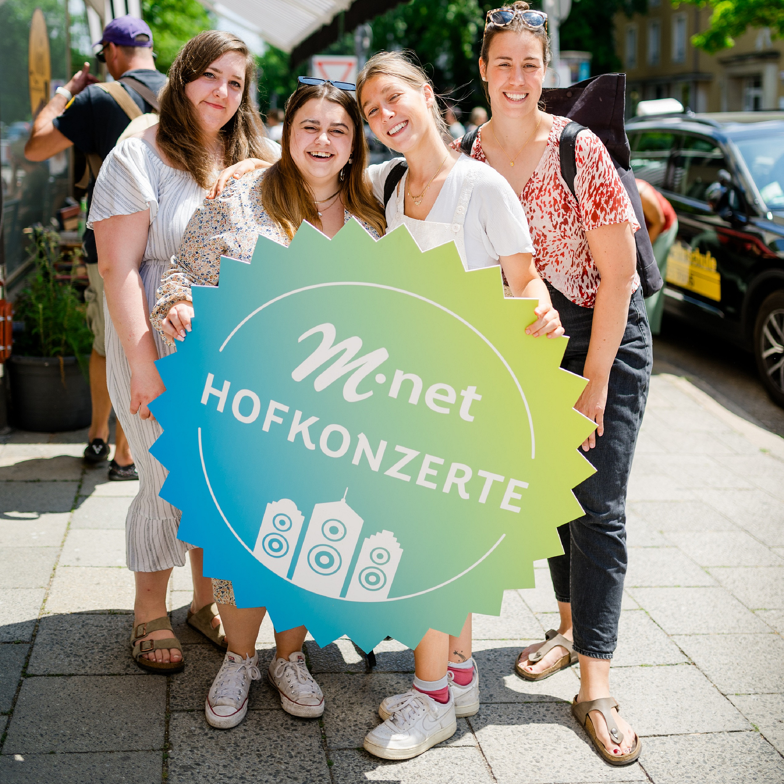 Vier Frauen stehen nebeneinander und halten in der Mitte ein Schild mir grün/blauem Farbverlauf auf dem in weißer Schrift "M-net Hofkonzerte" steht.
