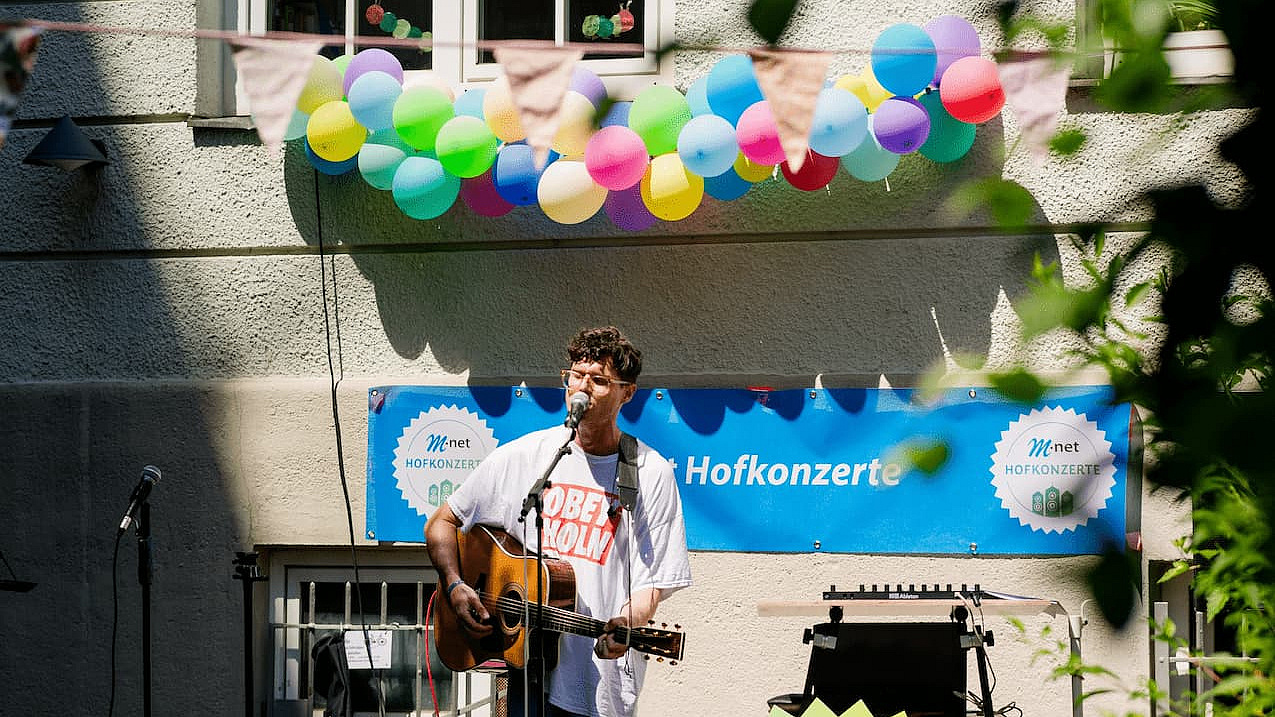 Sänger mit Gitarre vor einem M-net Plakat bei einem Hofkonzert