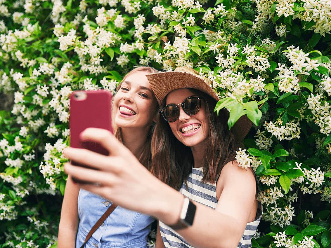 Zwei Frauen stehen vor einer blühenden Hecke und machen ein Bild von sich.