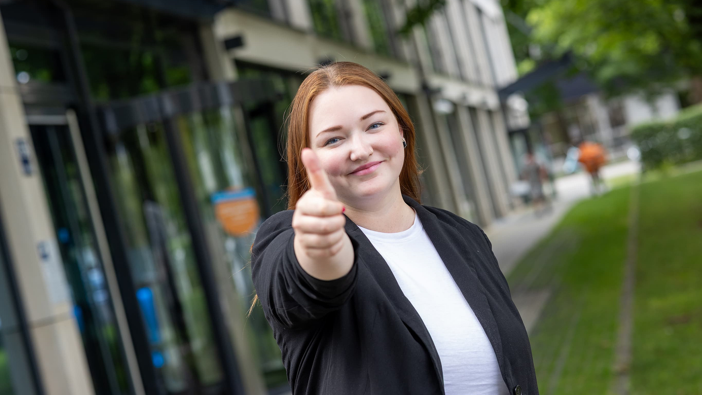Eine M-net Mitarbeiterin befindet sich vor dem Firmensitz im Außenbereich und zeigt den Daumen hoch 