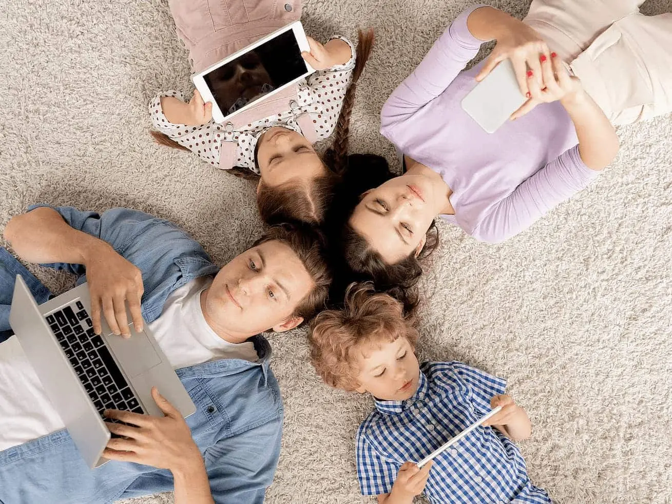Eine Familie liegt gemeinsam auf dem Teppich, jedes Mitglied hat ein Endgerät in der Hand, entweder Laptop, Handy oder Tablet
