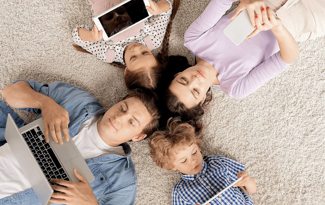 Vier Familienmitglieder auf dem Teppich liegend, alle haben je ein elektronisches Endgerät in der Hand