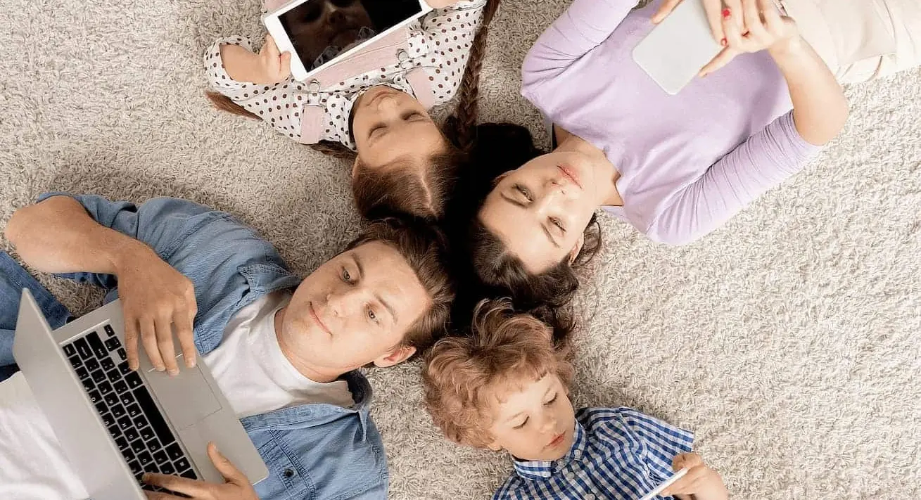 Eine Familie liegt gemeinsam auf dem Teppich, Ansicht von oben. Alle haben jeweils ein Tablet, Mobiltelefon oder einen Laptop in der Hand