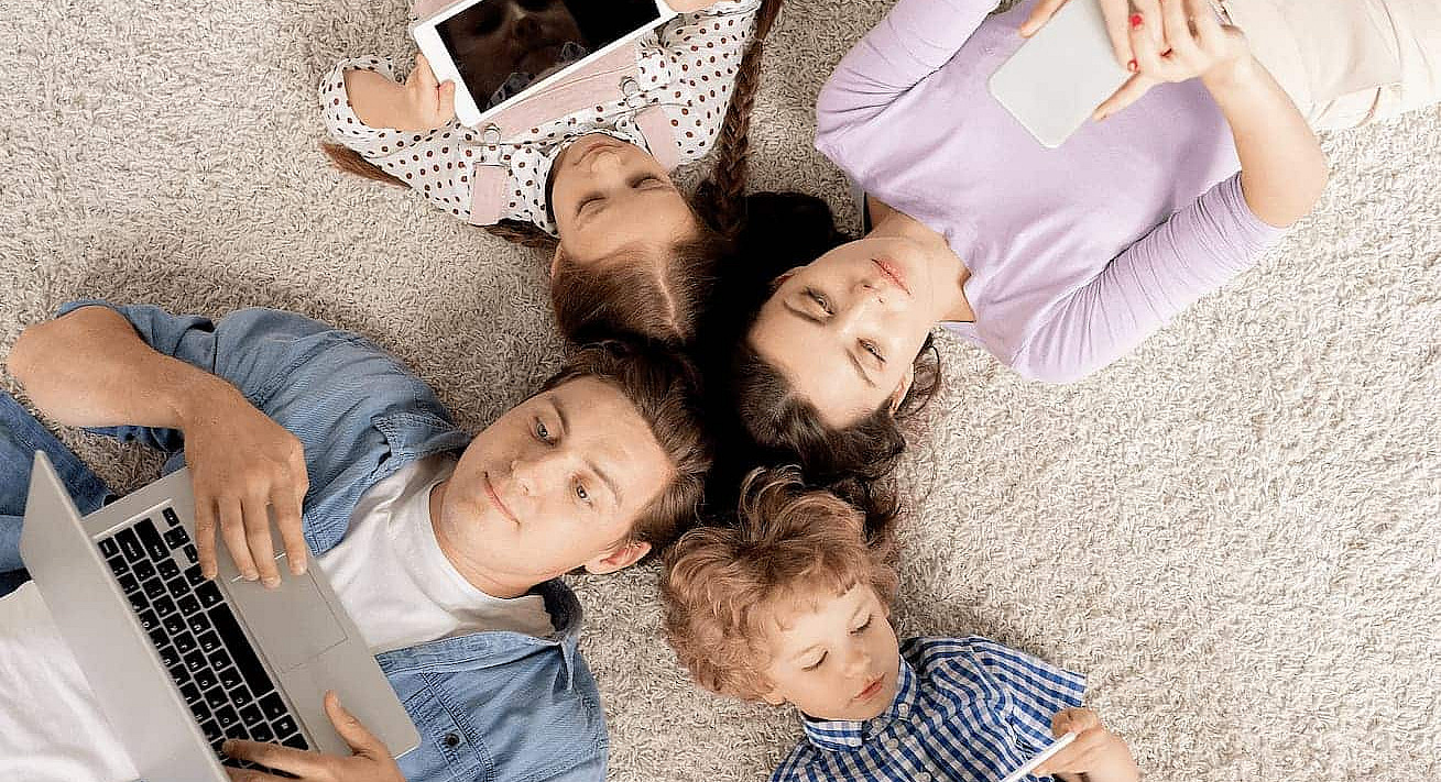 Eine Familie liegt gemeinsam auf dem Teppich, Ansicht von oben. Alle haben jeweils ein Tablet, Mobiltelefon oder einen Laptop in der Hand