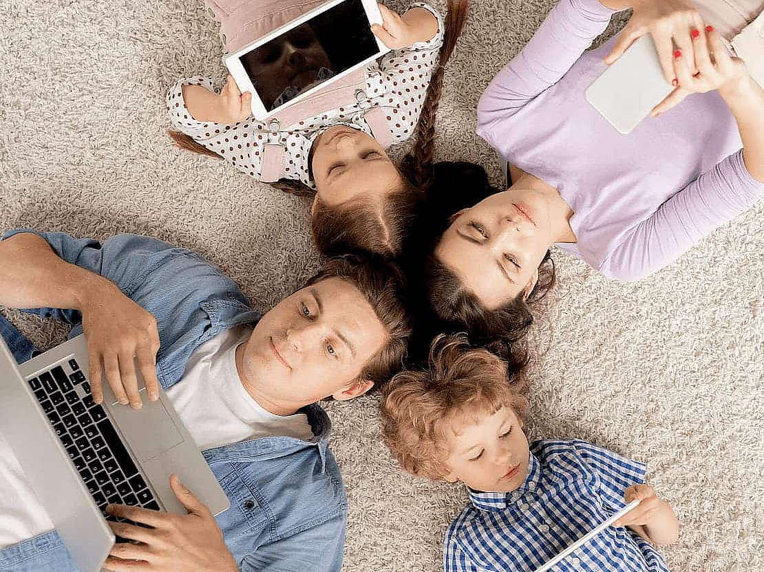 Vier Familienmitglieder auf dem Boden liegend, alle haben ein mobiles Endgerät in der Hand