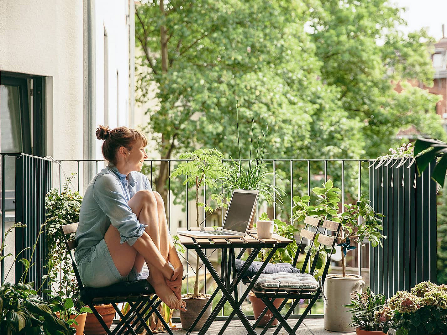Frau sitzt auf einem grün bepflanzten Balkon. Vor Ihr steht auf einem Balkontisch ihr Laptop und eine Kaffeetasse.