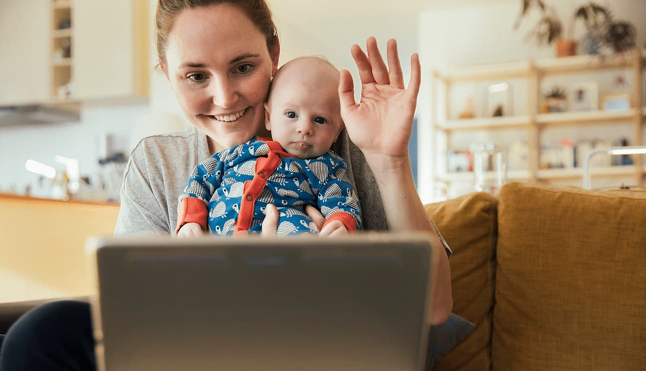 Eine Mutter sitzt winkend mit ihrem Baby im Arm vor einem Tablet