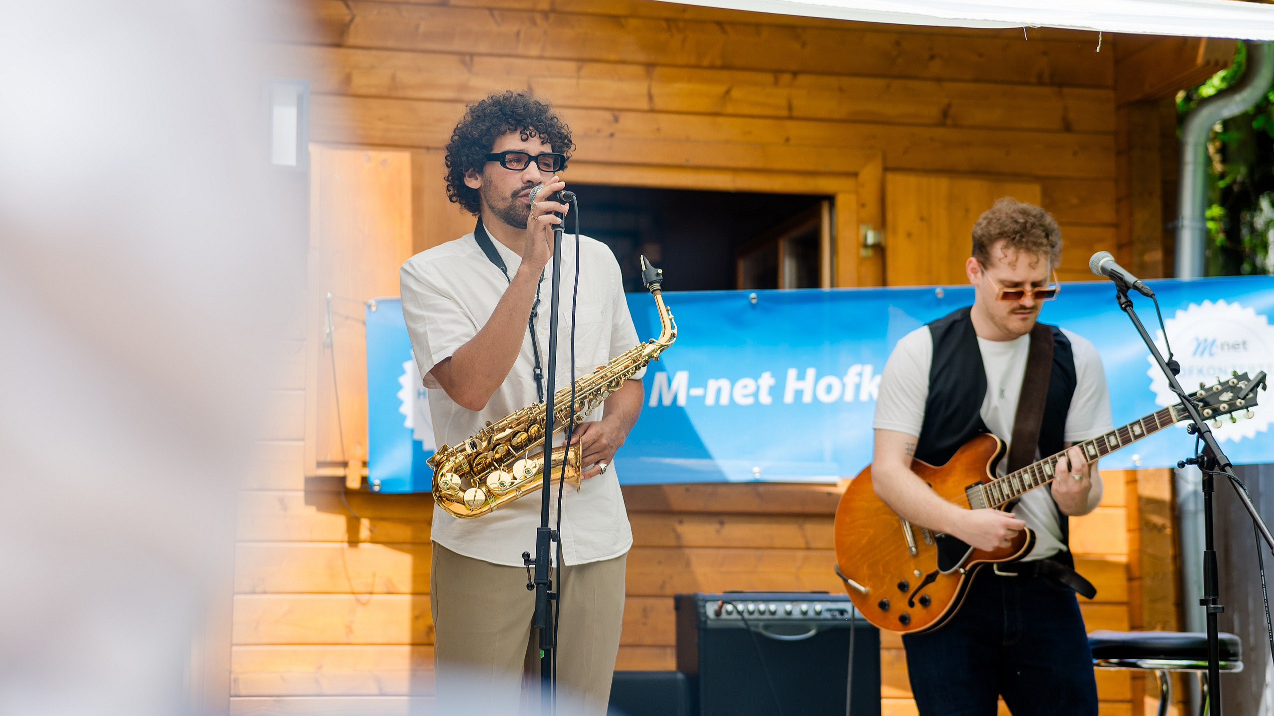 Zwei junge Männer stehen in einem Hinterhof eines M-net Hofkonzerts. Der linke Mann hält ein Mikrofon in der Hand und hat sein Saxofon umhängen. Der rechte Mann spielt Gitarre und hat ebenfalls ein Mikrofon vor sich stehen.