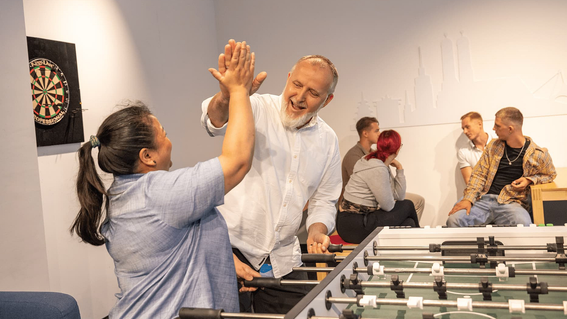 Kollegen im M-net Gemeinschaftsraum, im Vordergrund am Kicker freuen sich zwei Kollegen und klatschen ab