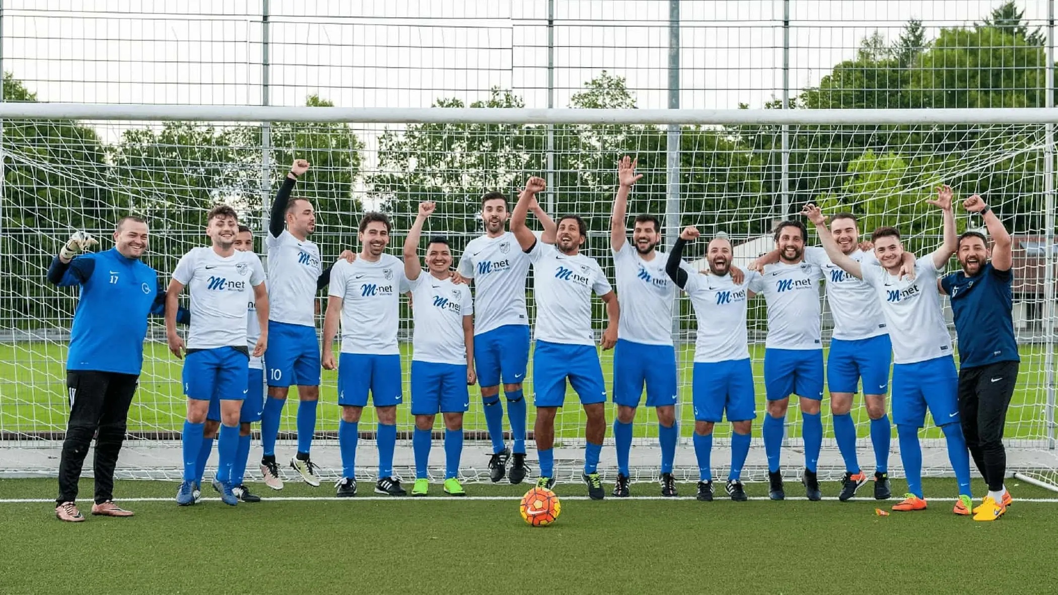 Eine Fußballmannschaft steht in einer Reihe auf dem grünen Spielfeld vor dem Tor. Vor Ihnen liegt ein bunter Fußball.