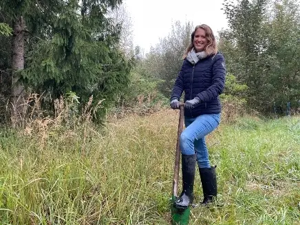 Eine blonde Frau mit langen Haaren und blauer Jeans steht an einem Waldstück auf einer Wiese. Sie lehnt sich auf einen Spaten.