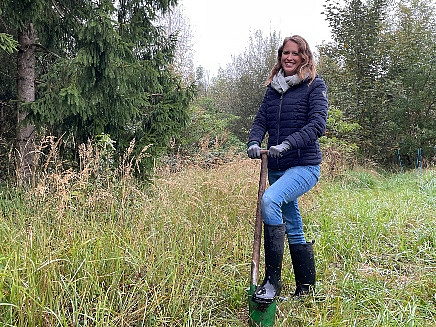 Eine blonde Frau mit langen Haaren und blauer Jeans steht an einem Waldstück auf einer Wiese. Sie lehnt sich auf einen Spaten.